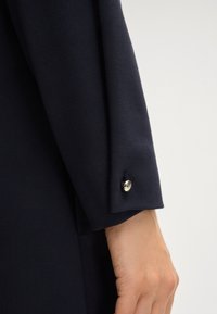 Marineblauwe langemouwen top van een gestructureerde stof. Heeft een knoopdetail bij de manchetten met een zilveren afwerking. Close-up van de mouw en hand.