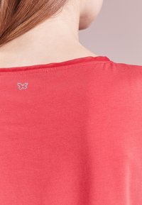 Camicia rossa con una texture liscia e un collo rotondo, caratterizzata da un piccolo design di farfalla di cristallo sul retro.