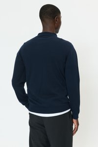 Mörkblå långärmad tröja med subtil struktur och ribbade muddar, visad bakifrån, bärs över en vit skjorta.