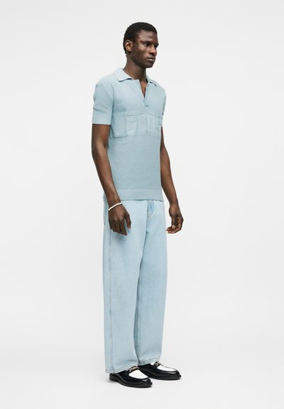 Άνδρας που φοράει ανοιχτό μπλε πόλο με κοντά μανίκια, ανοιχτό μπλε τζιν με φαρδιά παντελόνια, μαύρα και λευκά μοκασίνια και βραχιόλι με χάντρες, στέκεται μπροστά σε λευκό φόντο.