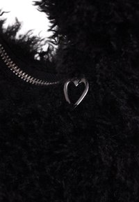 Textură pufoasă neagră cu închidere cu fermoar, având un accent mic în formă de inimă argintie atașat la material. Aspect moale și pufos.