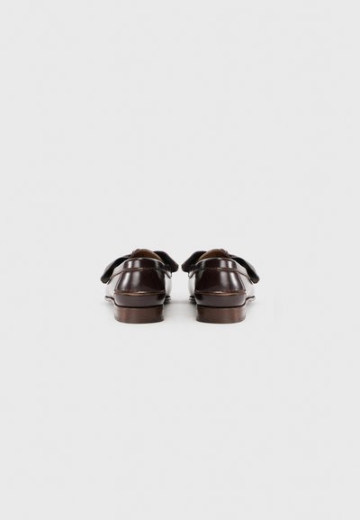 Pantofi din piele maro cu un finisaj neted, având vârful rotund și două fundițe decorative. Tălpile sunt plate și ușor ridicate.