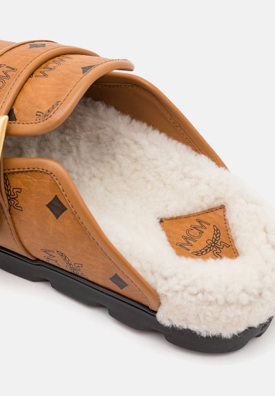 Pantoufle en cuir marron avec doublure blanche duveteuse, dotée d'une large lanière, de motifs en diamant et d'un patch logo MCM sur la semelle intérieure. Semelle extérieure noire.