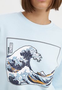 Sudadera de color azul claro con un diseño de ola en azul y blanco, con barcos naranjas y contorno negro. Tela suave con cuello de canalé.
