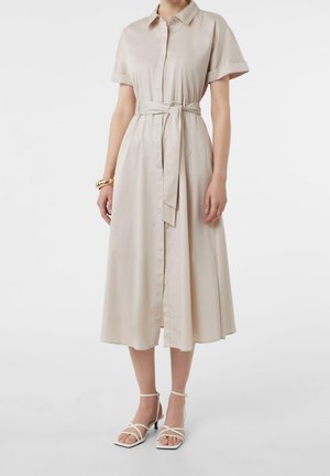 Robe chemise beige à manches courtes avec col, taille ceinturée, longueur midi, associée à des sandales blanches à talons avec brides et un bracelet en or.