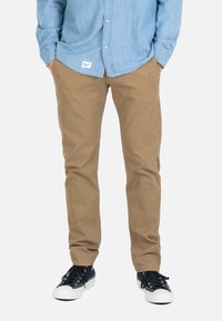 Hombre con pantalones caqui, camisa azul claro abotonada y zapatillas blancas y negras, de pie con las manos en los bolsillos.