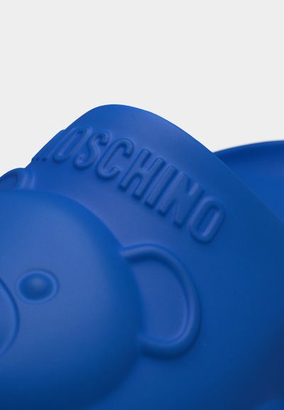 Μπλε αντικείμενο από καουτσούκ με ανάγλυφο πρόσωπο αρκούδας και ανάγλυφη επιγραφή "MOSCHINO" σε κυρτή επιφάνεια.
