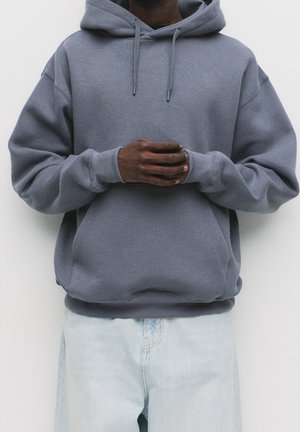 Hombre vestido con una sudadera gris con capucha, con las manos entrelazadas al frente, combinado con vaqueros de mezclilla azul claro sobre un fondo liso.