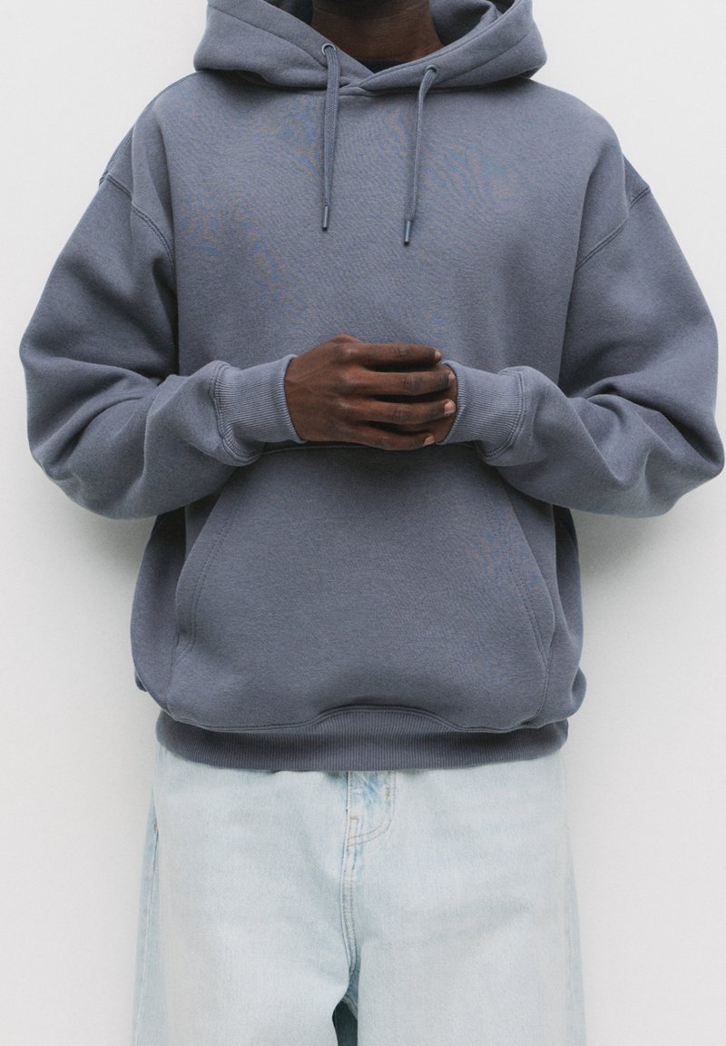 Homme portant un sweat à capuche gris, les mains jointes devant, associé à un jean en denim bleu clair sur un fond uni.