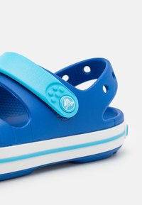 Sanda azul de goma con una correa ancha con un acento azul claro, recortes circulares para ventilación y una suela blanca y azul a rayas.