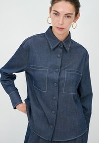 Camicia in denim blu scuro con bottoni, caratterizzata da cuciture bianche a contrasto, due tasche frontali e un design con colletto classico.