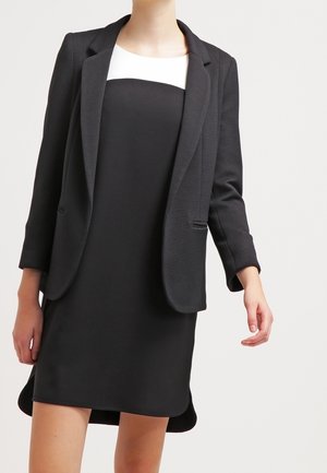 Frau trägt ein schwarz-weißes ärmelloses Kleid kombiniert mit einem schwarzen, strukturierten Blazer und steht vor einem einfarbigen hellen Hintergrund.