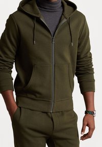 Sudadera con cremallera en color verde oliva, con un cierre frontal, bolsillos amplios y cordones. Puños y dobladillo acanalados, hecha de una tela suave con una textura lisa.