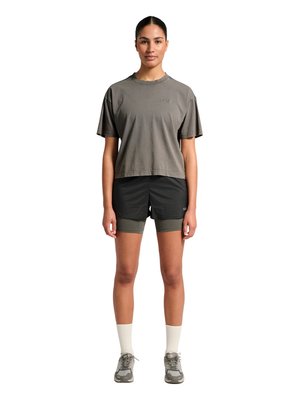 Kvinde stående med front mod kamera iført grå kortærmet skjorte, sorte shorts over olivengrønne cykelshorts, hvide sokker og grå sneakers på hvid baggrund.