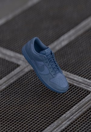 Blue Nike sneaker with laces resting on a metal grid surface with diagonal lines in an outdoor setting.