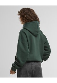 Mørkegrønn hettegenser med en avslappet passform, ribbestrikkede mansjetter og en myk tekstur. Kombinert med mørkegrå jeans, sett fra baksiden.