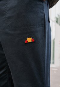 Námořnické modré kalhoty s hladkou texturou, které mají malé vyšívané logo: polokruh v žluté a červené barvě na přední kapse.