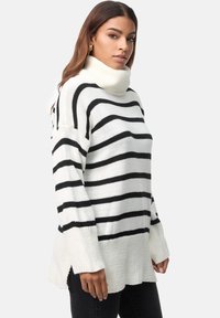 Pull à col roulé rayé en blanc et noir, présentant une coupe ample, des poignets côtelés, des fentes latérales et un motif tricoté texturé.