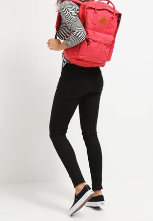 Personne portant un pantalon noir, une chemise à manches longues rayée, des baskets noires, et portant un sac à dos rouge vif sur le dos.