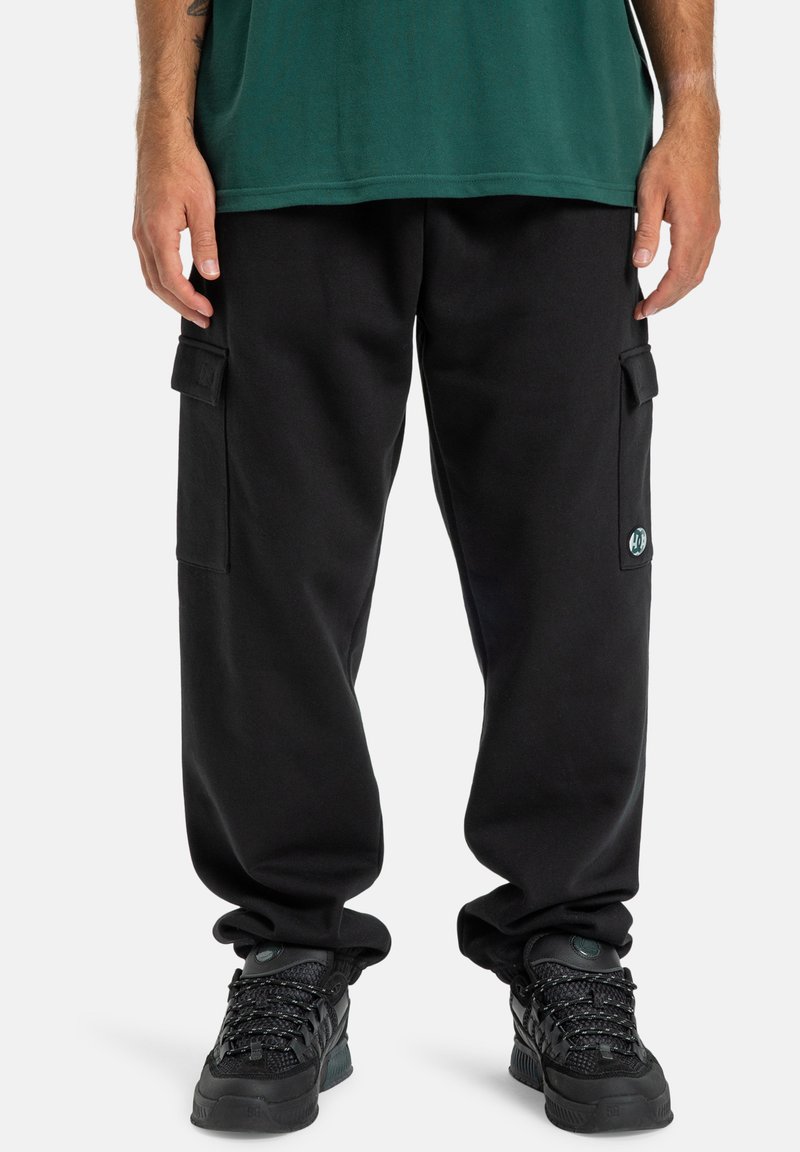 Schwarze Cargo-Hosen mit seitlichen Taschen, aus weichem Stoff gefertigt. Mit lockerer Passform und elastischen Bündchen an den Knöcheln, kombiniert mit schwarzen Sneakers.