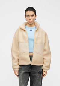 Veste en polaire beige à la texture douce, fermeture éclair, et panneau avant crème. Associée à un haut ajusté bleu clair et un jean foncé.