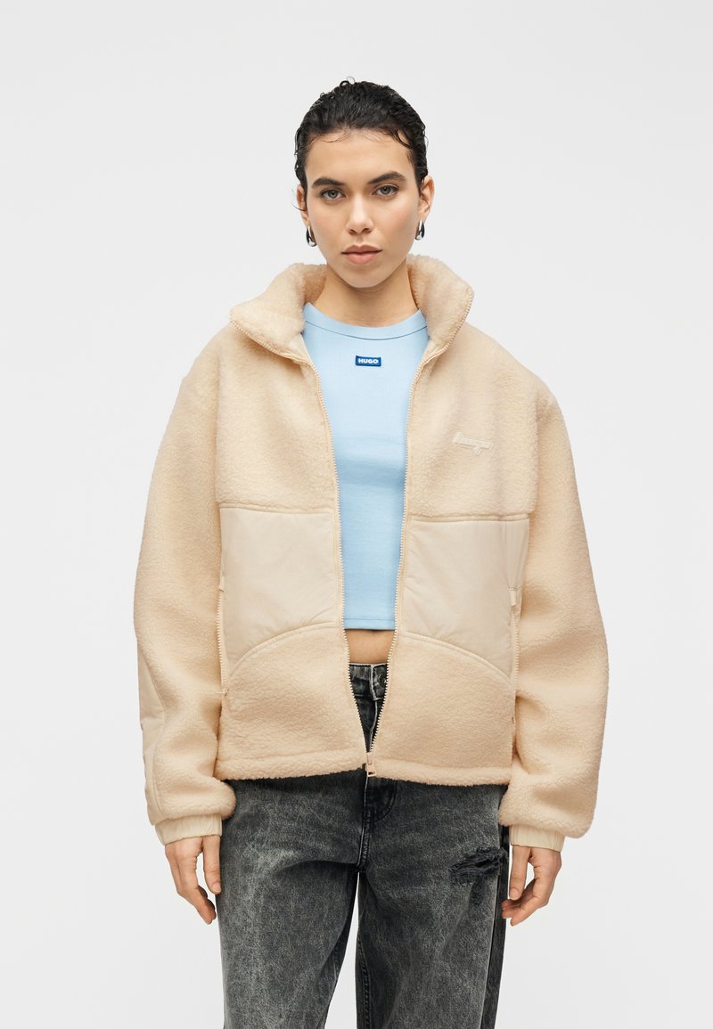 Veste en polaire beige à la texture douce, fermeture éclair, et panneau avant crème. Associée à un haut ajusté bleu clair et un jean foncé.