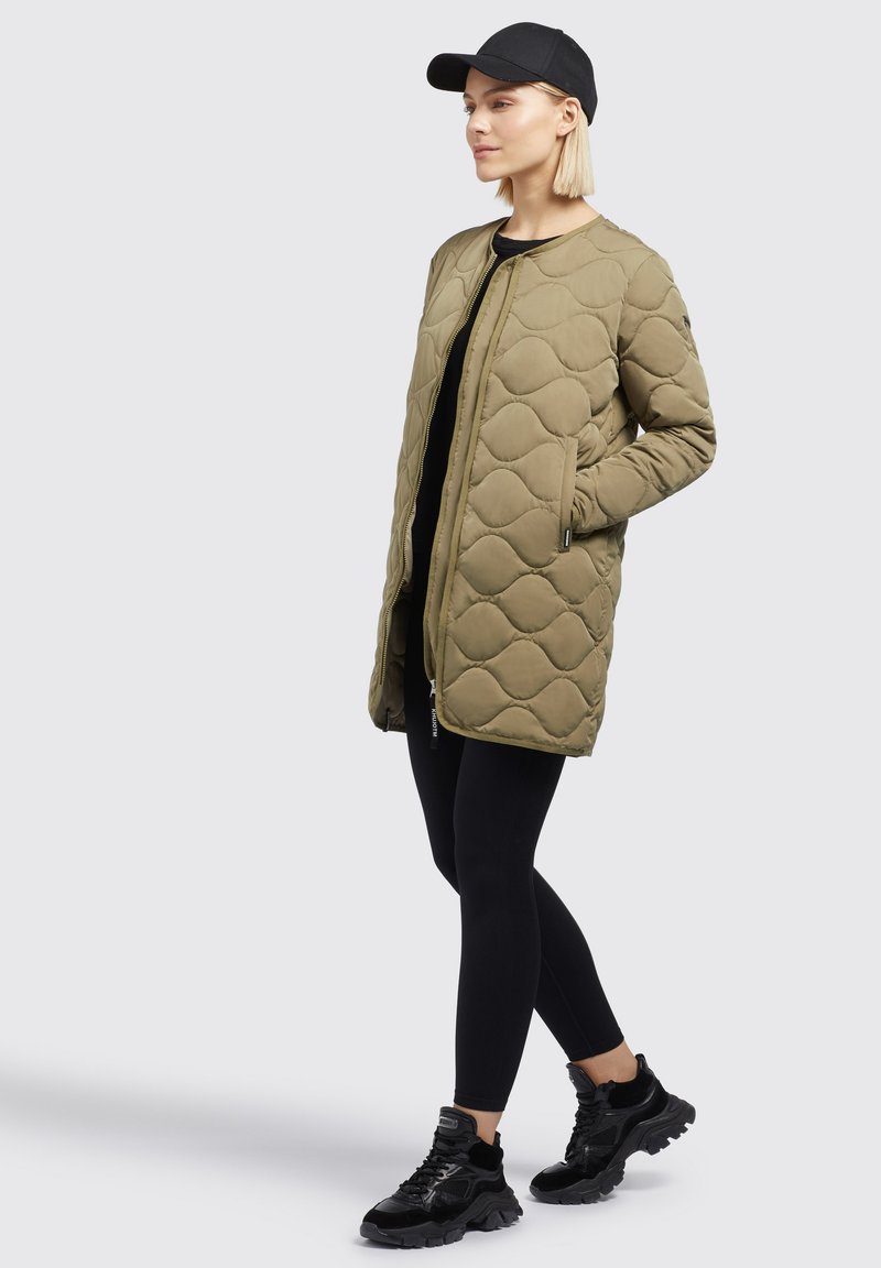 Gestepptes olivgrünes langes Jacket mit Reißverschluss und Seitentaschen, getragen über schwarzen Leggings und schwarzen Sneakers, ergänzt durch eine schwarze Mütze.