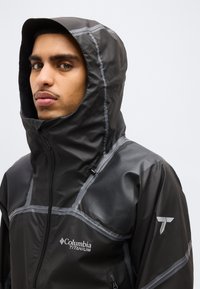 Άνδρας που φοράει μαύρο αδιάβροχο μπουφάν Columbia Titanium με κουκούλα και αντανακλαστικές ραφές, κοιτάζοντας ελαφρώς προς την πλευρά σε απλό φόντο.
