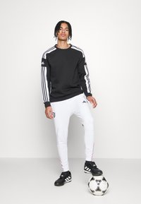 Schwarzer Sweatshirt mit weißen Streifen, weiße Jogginghosen mit einem Logo, schwarze Fußballschuhe und ein Fußball. Glatter Stoff und sportliche Passform.