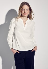 Vit blus med V-urringning och volangkant, med långa ärmar med texturerade detaljer. Kombinerad med marinblå byxor.
