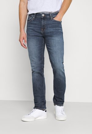 Mężczyzna noszący dopasowane niebieskie jeansy i białe trampki, stojący z jedną ręką w kieszeni na jednolitym jasnym tle.