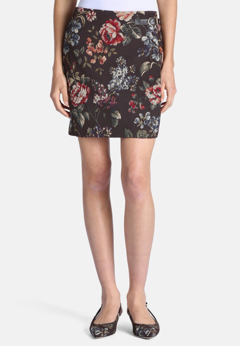 Femme portant une jupe courte noire à fleurs avec une petite boucle latérale et des chaussures plates fleuries assorties, debout devant un fond uni.