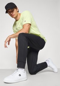 Helles grünes T-Shirt mit gesticktem Logo, schwarze Jogginghose und weiße Sneakers. Das Ensemble hat eine legere, sportliche Passform.