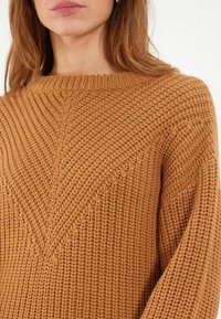 Pull en tricot ample de couleur marron chaud, avec un col rond et un détail en V en relief sur le devant. Tissu doux et texturé.