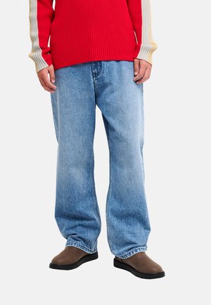 Personne portant un jean bleu clair ample, des chaussures marron, et un pull rouge avec des manches rayées beige et grises.