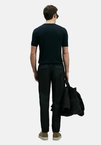 Zwarte ribgebreide trui met korte mouwen, gecombineerd met zwarte broeken. Man houdt een zwarte jas vast. Lichtbruine schoenen maken de outfit compleet.