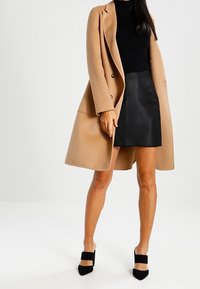 Cappotto cammello con bottoni doppi, dolcevita nera, gonna nera testurizzata e tacchi neri con cinturini, su uno sfondo neutro.