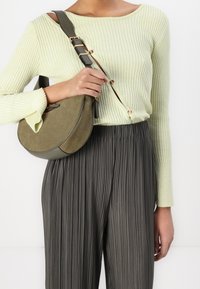 Donna con un top a coste verde chiaro con bottoni e pantaloni plissettati scuri, che tiene in mano una borsa a forma di mezzaluna in suede verde oliva.