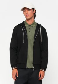 Felpa nera con zip, dotata di un cordoncino grigio e tasche laterali, indossata sopra una maglietta polo verde oliva a trama. Presenta polsini a righe.