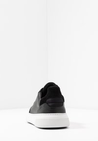 Svart lädersneaker med mockadetaljer, lågprofil design, vadderad krage och vit gummisula, med strukturerat grepp och sömnadsdetaljer.