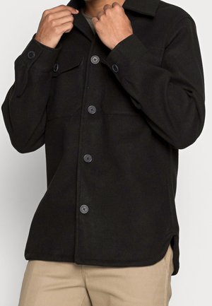 Mężczyzna poprawiający kołnierz czarnej wełnianej kurtki zapinanej na guziki z klapkowymi kieszeniami na wysokości klatki piersiowej, ubrany w beżowe spodnie.