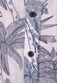 Valkoinen kangas, jossa on tummansininen trooppinen lehtikuvio, jossa on kaksi mustaa nappia pystysuorassa paitapieluksessa.