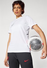 Biela športová tričko s krátkymi rukávmi a čiernym Nike swooshom. Tmavošedé nohavice s červeným logom Nike. Hráč drží čierno-biely futbalový loptu.