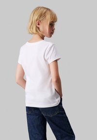 Camiseta blanca de algodón con mangas cortas, escote redondo y textura suave, llevada con jeans de mezclilla azules, mostrada desde la espalda.
