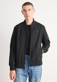 Black bomber jacket with a zipper front, ribbed cuffs and hem, and a subtle logo on the chest. Worn over a black shirt and light blue jeans.