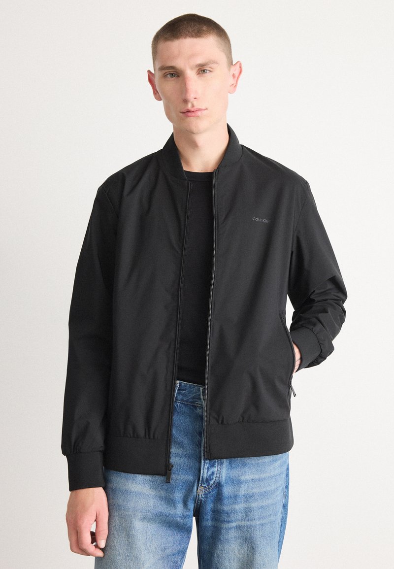 Black bomber jacket with a zipper front, ribbed cuffs and hem, and a subtle logo on the chest. Worn over a black shirt and light blue jeans.