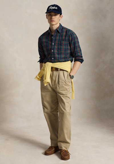 Jonge man met een groen geruit overhemd, beige broek, gele trui gestrikt rond de taille, bruine schoenen en een navy pet met "Ralph" logo, staand met handen in de zakken.
