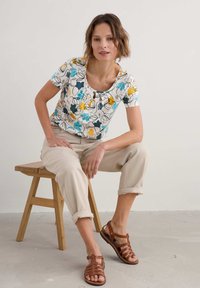 Bloemmotief top met blauwe, gele en zwarte bloemen op een witte achtergrond; ronde halslijn; gecombineerd met beige cropped broeken en bruine sandalen.