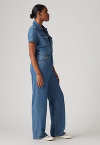 Lys blå denim jumpsuit med korte ermer, knappestenging foran og vide ben. Har frontlommer og en trent midje.
