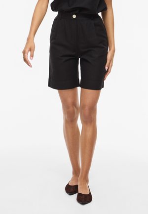 Osoba nosiaca čierne šortky s vysokým pásom a gombíkom, čierny top a hnedé ploché topánky, stojaca pred jednoliatym bielym pozadím.